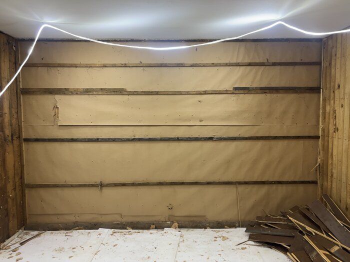 Rivet väggparti med borttagen tapet och exponerade brädor i ett rum under renovering. Brädrester ligger på golvet.