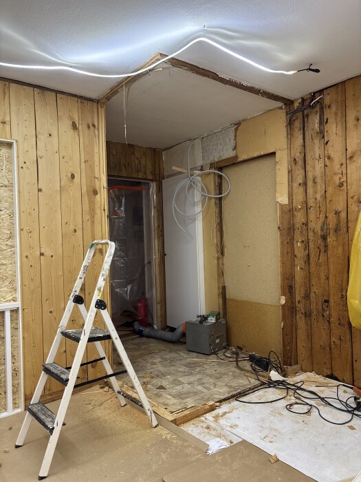 Rum under renovering med borttagen tapet, stege, sladdar och verktyg på golvet.