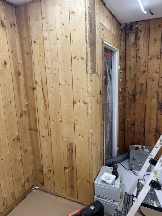 Rum under renovering med omålade träväggar, utrustning och stege. Väggar är delvis utan tapet. Verktyg och kablar syns på golvet.