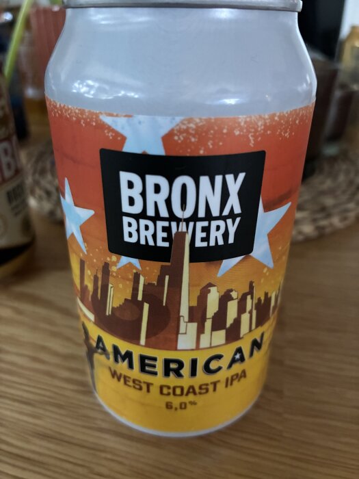 Burk med Bronx Brewery American West Coast IPA, 6 %; gul etikett med orange och vita stjärnor samt stadslandskap, på ett träbord.