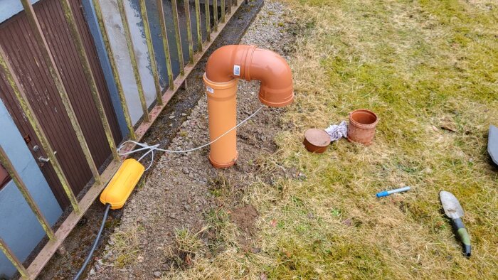 Orange rörfläkt installerad vid dräneringens inspektionsbrunn, kopplad med en vit kabel på grusig mark bredvid en gräsmatta och staket.