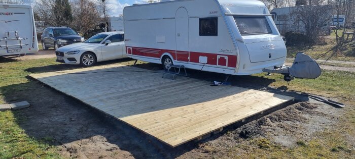 Husvagn parkerad bredvid en bil på en nybyggd träplattform i en trädgård.