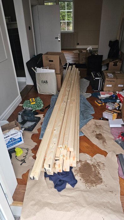 Trähögar och byggmaterial på golvet i ett rum under byggnation, omgivna av verktyg och kartonger.