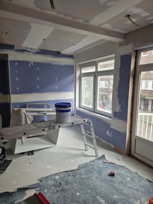 Renoverande kök med tvådelat fönster, stege, vit färgburk, och oskyddade väggar i förberedelse för installation av köksskåp.