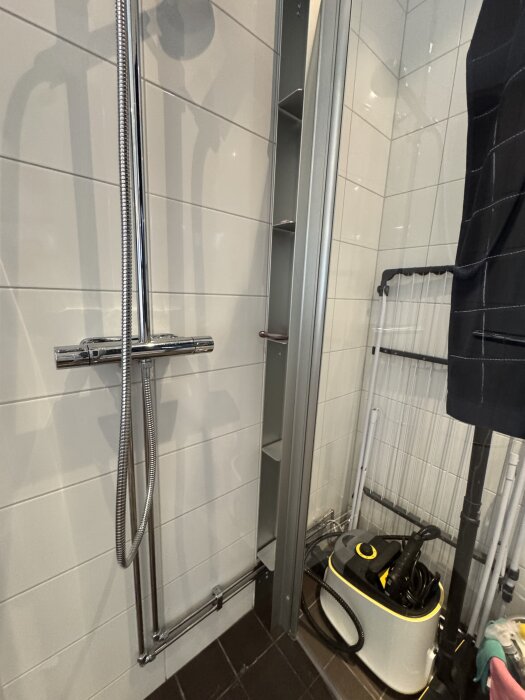 Duschhörna med vitt kakel och en modern duschinstallation. I hörnet finns en städmaskin och ett hopfällbart torkställ.