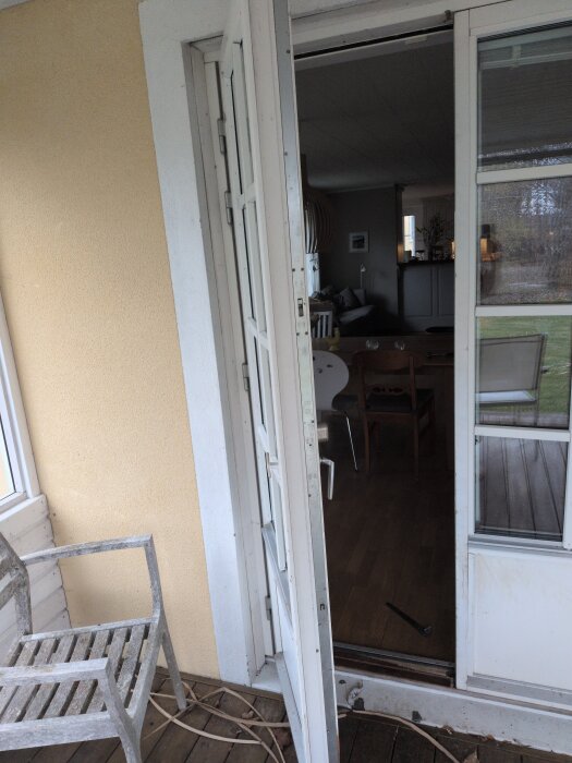 Öppen altandörr med synligt låsmejsel och trädgårdsstol bredvid, insidan av huset syns i bakgrunden.