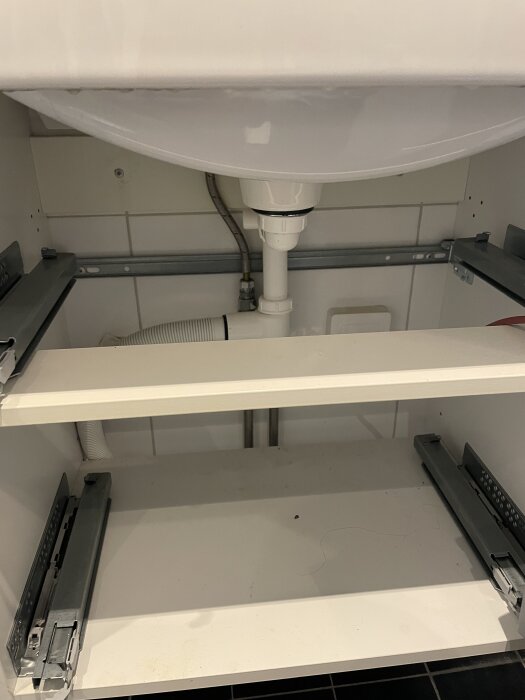 Handfat med synligt rör och avloppsinstallation i ett badrumsskåp. Hyllplan och skenor för lådor synliga under handfatet.
