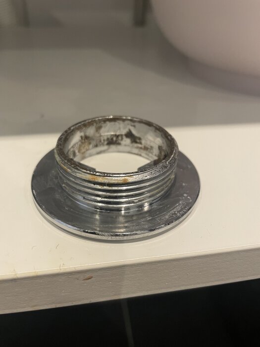 Metallgänga och fläns, troligen från VVS, placerad på vit yta. Gängans yta visar tecken på rost eller slitage.