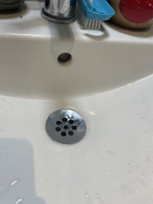 Vitt handfat med silverfärgad avloppssil, en tandborste i blå hållare och en tvålpump vid sidan. Vattendroppar syns på ytan.