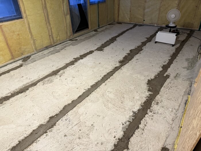 Betonggolv med spår fyllda med cement i ett rum som håller på att renoveras, en fläkt och element i hörnet.