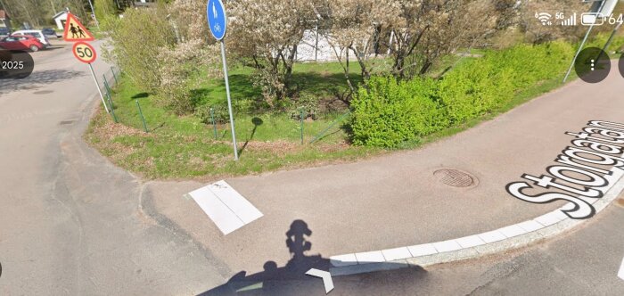 En vägkorsning med trafikskyltar, trottoarkant och vägmarkeringar, omgiven av grönska. Vägskylt visar gående och cyklister.