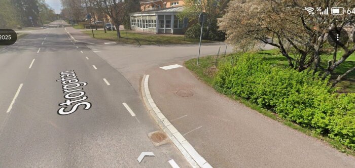 Gatukorsning där en huvudgata svänger av mot en mindre väg, omgiven av träd och buskar med trottoar utmed vägen.