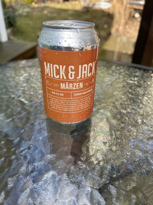 En ölburk med etikett "Mick & Jack Märzen" står på ett glasbord utomhus, täckt av vattendroppar.