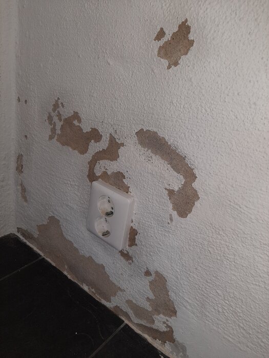 Vägg med avskavd färg och blottad murputs ovanför ett dubbeluttag.