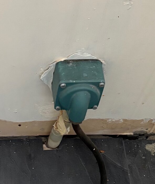 Grön elbox monterad på en vägg, med svart kabel som går ner mot ett mörkt golv, omgiven av reparerat och målat väggmaterial.