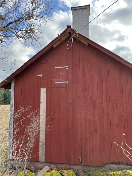 Rött träskjul med eternittak, tegelstensskorsten och korta reglar under takfoten nära gavelspetsen sett mot en molnig himmel.