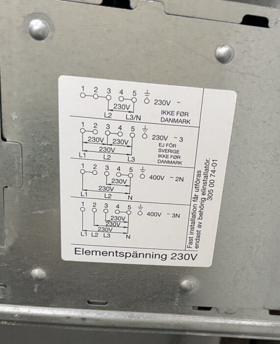 Etikett på metallpanel som visar kopplingsscheman för elanslutningar med olika spänningar, märkt "Elementsättning 230V", med varningstext.