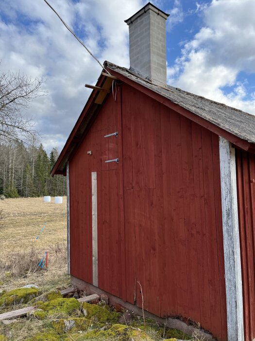 Rött hus med eternittak och skorsten. Kortare reglar syns under takfoten nära gavelspetsen. Skog i bakgrunden och molnig himmel.