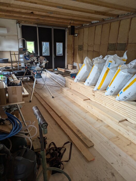 Verkstad under renovering med staplat virke på golvet, såg och diverse verktyg, insäktade väggar och svart dörr i bakgrunden.