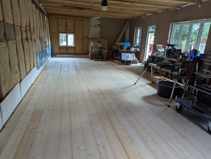 Nyinstallerat massivt trägolv i verkstad med verktyg och byggmaterial längs väggarna, stora fönster ger gott om ljus.