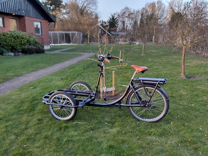 Lådcykel utan låda med applicerad underredsmassa parkeras på gräsmatta vid villa, synliga skärmar och träramar i bakgrunden.