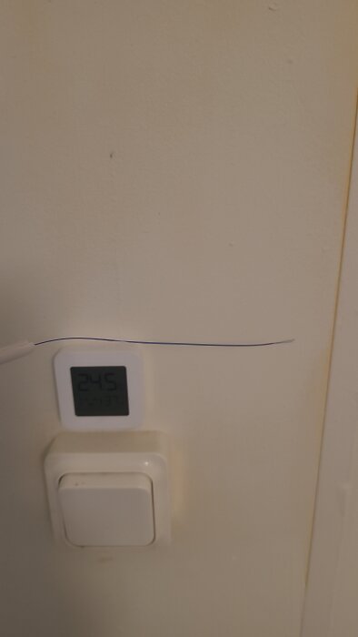 Termometer visar 24,5°C på väggen bredvid en ljusströmbrytare, med en tunn kabel som löper över dem.