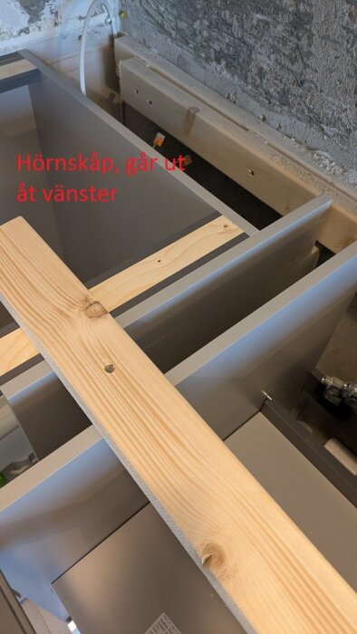 Hörnmonterat LeMans-skåp med synlig träplanka och text "Hörnskåp, går ut åt vänster" på oslipad vägg i bakgrunden.