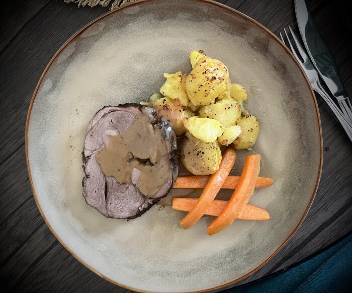 Lammstek med sås, smörslungade morötter och ugnsstekt potatis, serverat på en beige tallrik med gaffel och kniv vid sidan.