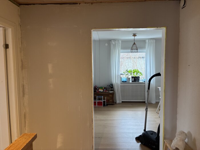 En nyligen bredspacklad hallvägg syns, med en dammsugare i ett ljust rum i bakgrunden, samt ett fönster med gardiner och växter.