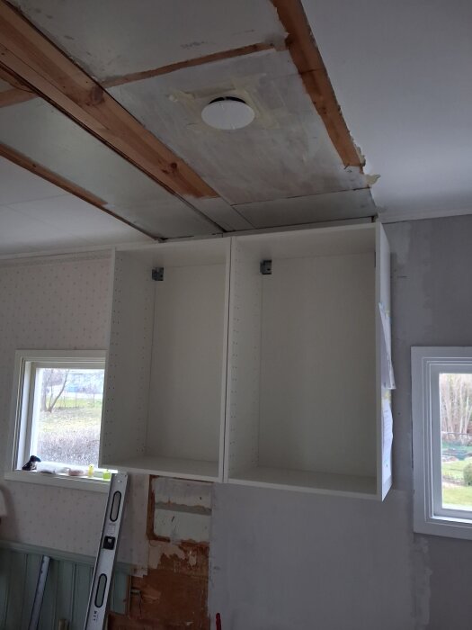 Öppna väggskåp under tak med nivåskillnad; delar av taket saknar panel och masonite är synlig. En fönsteröppning syns vid sidan.