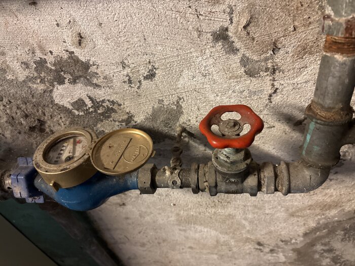 Old water meter with a brass cover next to a red valve on a corroded pipe against a rough, painted wall.