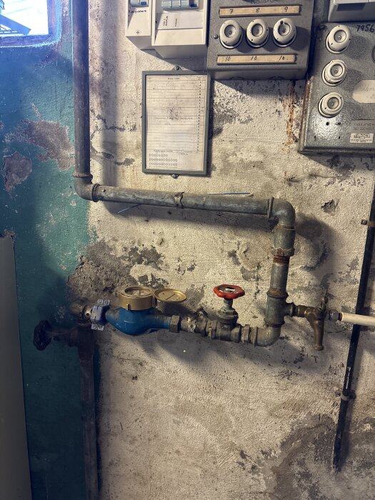 Old plumbing pipes with gauges and valves on a worn concrete wall, next to electrical switches and a handwritten note.