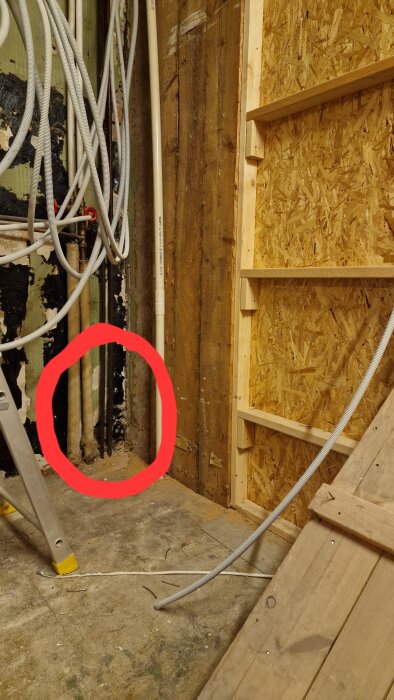 Byggarbetsplats med exponerade rör och kablar mot en trävägg i ett rum under renovering. En stege står i hörnet, och några rör är markerade med en röd cirkel.