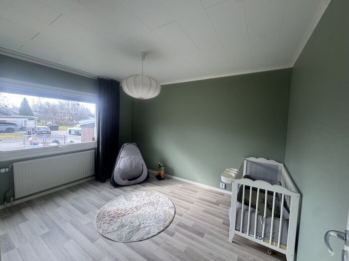 Barnrum med ljusgrönt väggfärg, spjälsäng, runt matta och tält nära stort fönster med utsikt över en gata.