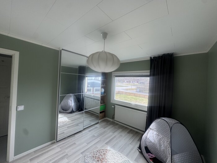 Barnrum med gröna väggar och parkettgolv, stort fönster med gardin, spegelskåp och lektält i hörnet, rummet nästan färdigt.