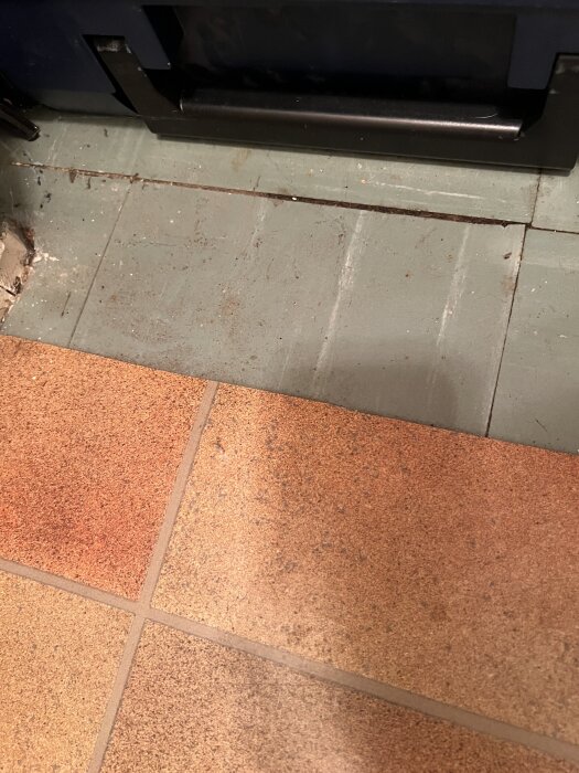 Grönt golv med spår av borttagen plastmatta och lim ovanpå ursprungligt trägolv, delvis synligt bredvid brunt klinkergolv.