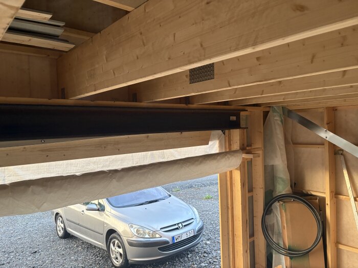 Garageinteriör med synliga träbjälkar och en stålbalk ovanför porten. Utanför garaget syns en bil parkerad på grus.