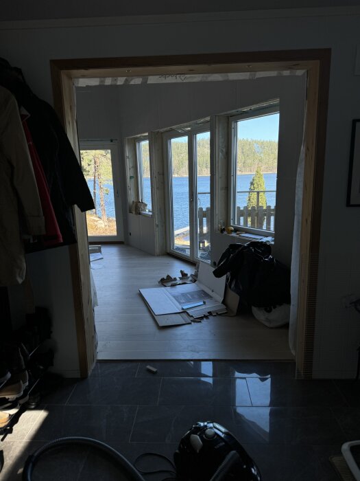 Ett delvis möblerat rum med nygjort golv, skjutdörrar mot utsikt över en sjö utanför. Byggmaterial syns på golvet.