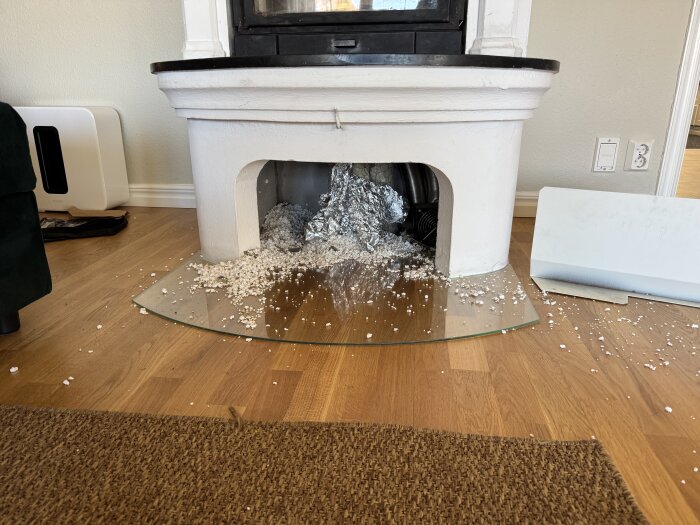 Cellplast spritt framför en vit kamin på ett trägolv, med glasunderlägg och silverfärgat material inuti kaminen.