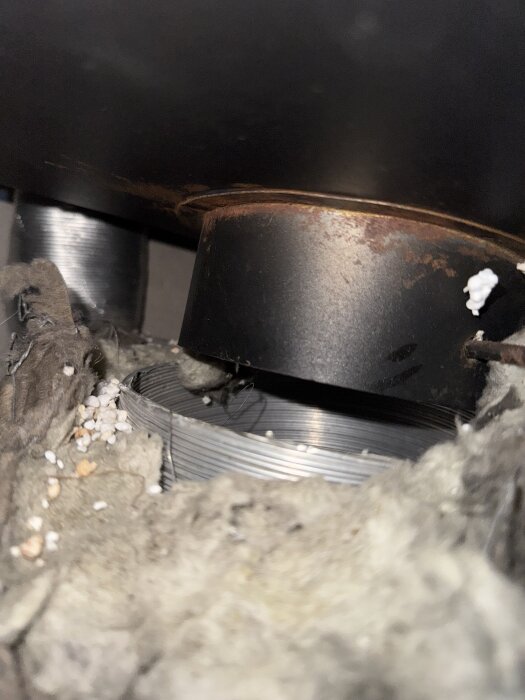 Rostig kaminanslutning med ventilationsrör, omgiven av isoleringsmaterial och små bitar av cellplast.