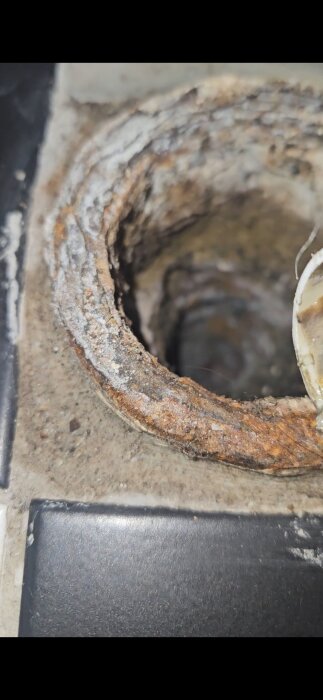Gammalt gjutjärnsavloppsrör i golv, med rost och slitage synligt på insidan av röret. Avsett för handfatsdränering.