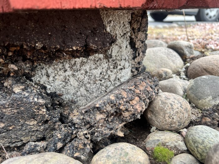 Vittrat mörkt skyddsmaterial runt en betongplatta på ett hus från 1973, med småsten runt om, ses på bilden.