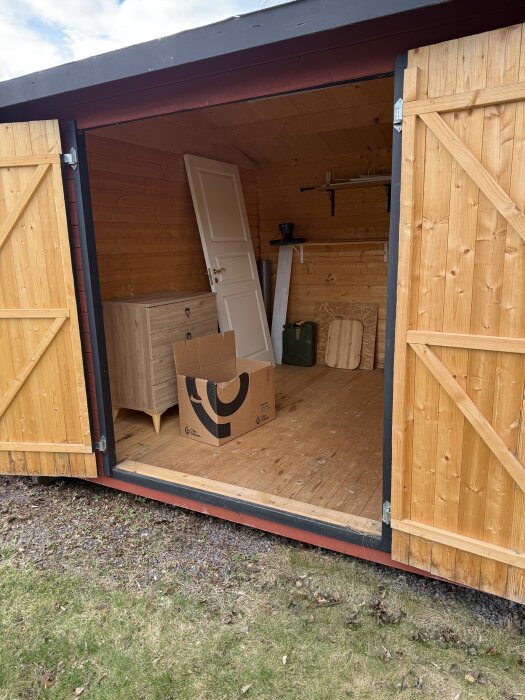Öppet förråd med trädörrar, trägolv, vit dörr lutad mot väggen, hylla och kartong. Trägolv och väggpanel i ljus träfärg.