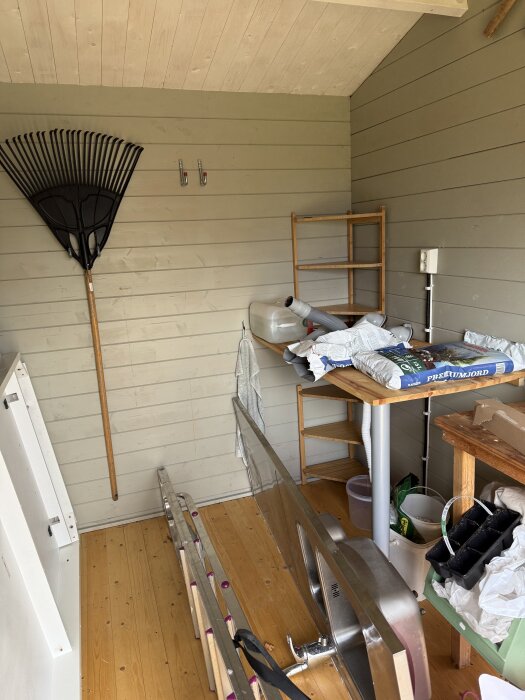 Inre del av gammalt förråd med träpanel på väggarna, olika föremål och verktyg. Förrådet planeras att renoveras till en gäststuga.
