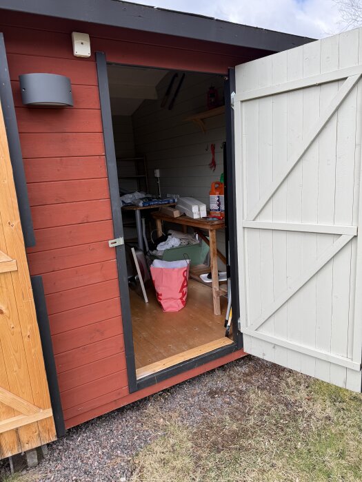 Rött förråd med öppen dörr, inuti syns verktyg, hyllor och renoveringsmaterial, grått paneltak och ljus trädetalj på dörren.