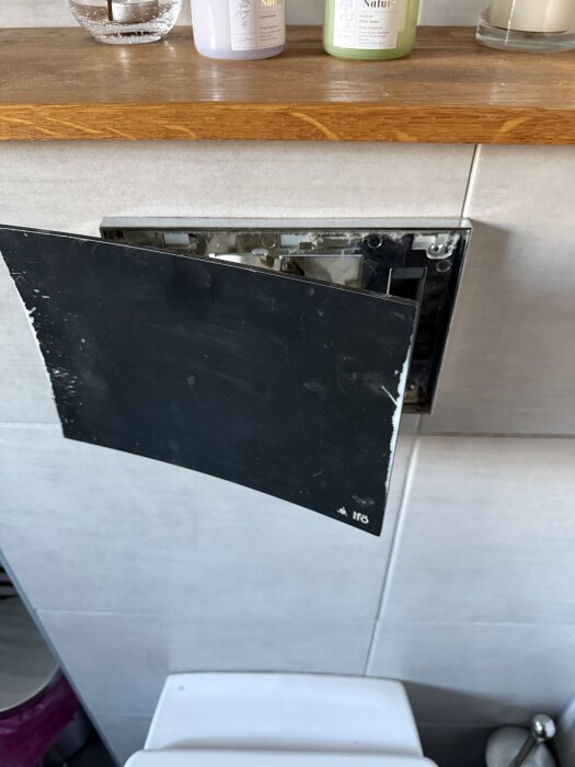 Svart frontpanel från spolknapp på Ifö toalett är delvis borttagen, med en hylla med flaskor ovanför.