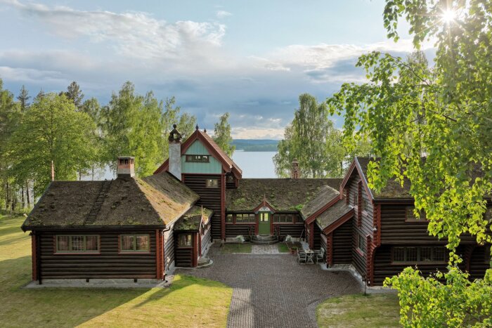 Stor timmerstuga med grästak vid en sjö, omgiven av gröna träd och växter, med vy över vattnet och himlen i Falun.