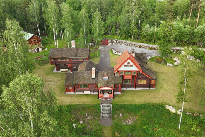 Stort rött trähus med grönt tak och detaljer, omgiven av grön skog och gräsmatta i Falun.