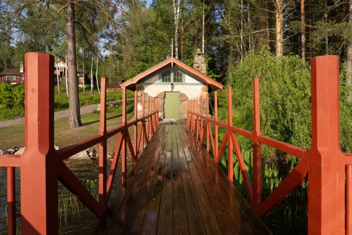 Röd träbro som leder till en liten byggnad med grön dörr, omgiven av träd och buskar, i ett solljust landskap.