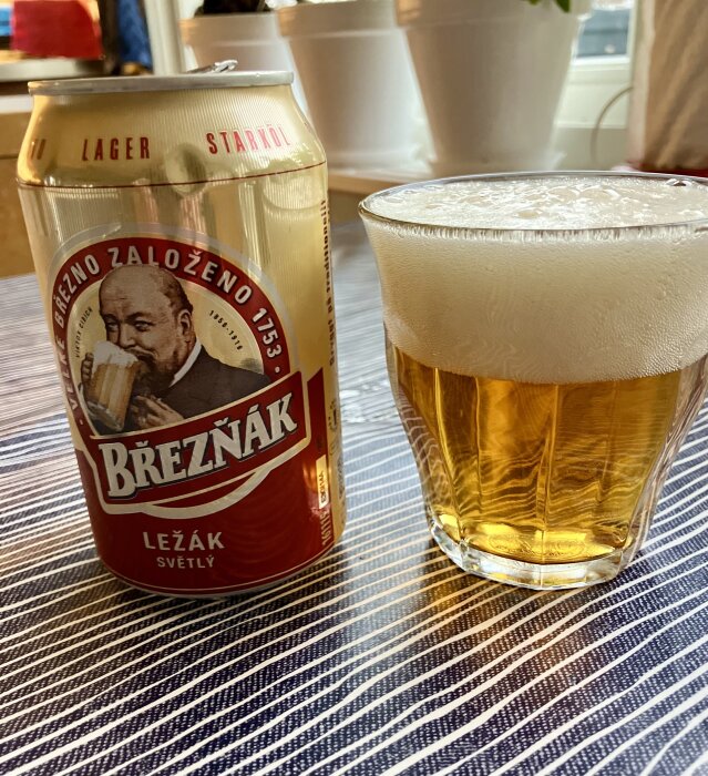 En burk Březňák-bärs och ett glas öl med skum på en randig yta.